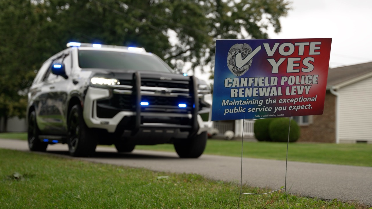 A Vote Yes yard sign for the Canfield police renewal levy on a front lawn, with a police SUV blurred in the background